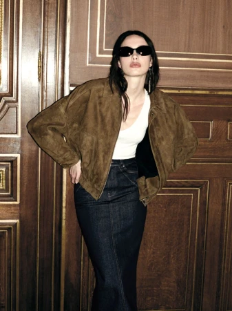 Model wearing dark sunglasses, brown suede jacket, and long denim skirt leaning against a dark wood wall