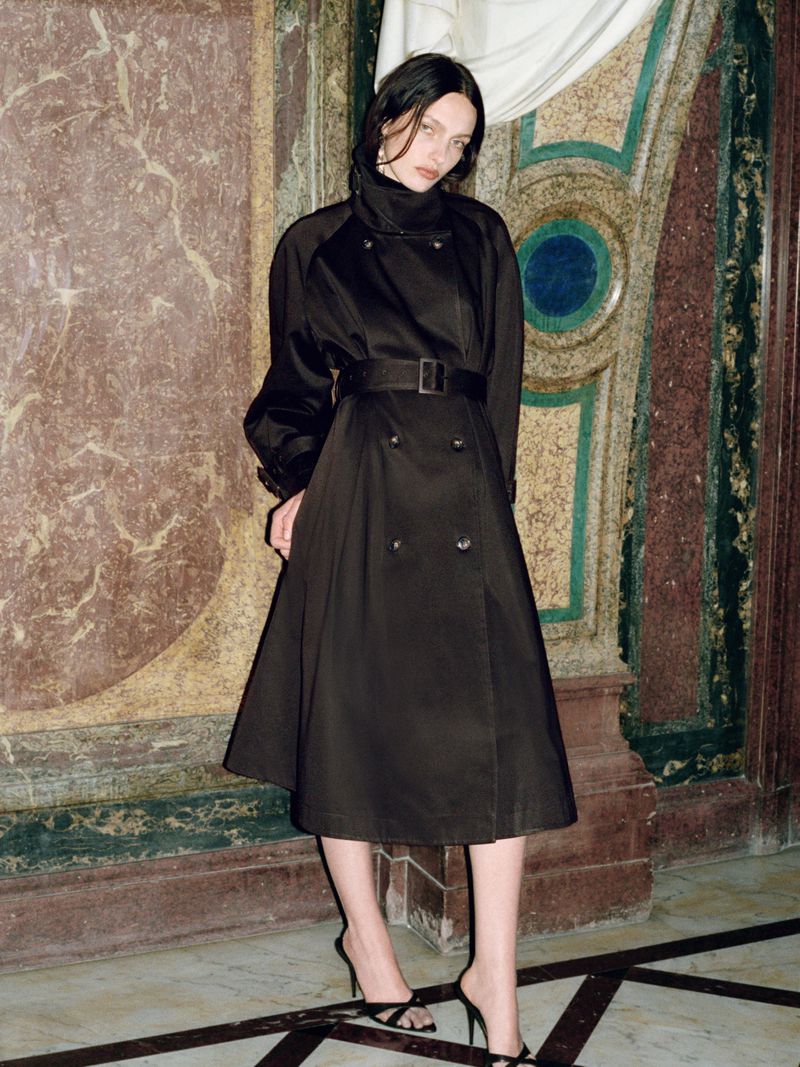 Model in a black belted trench coat standing before ornate classical stone pillars and marble walls.