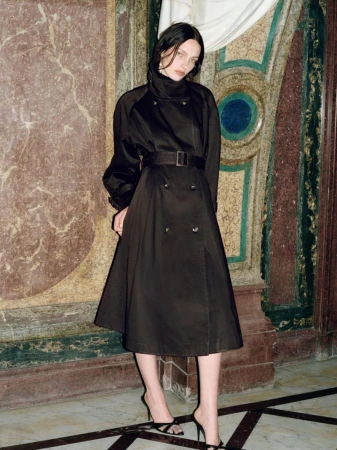 Model in a black belted trench coat standing before ornate classical stone pillars and marble walls.