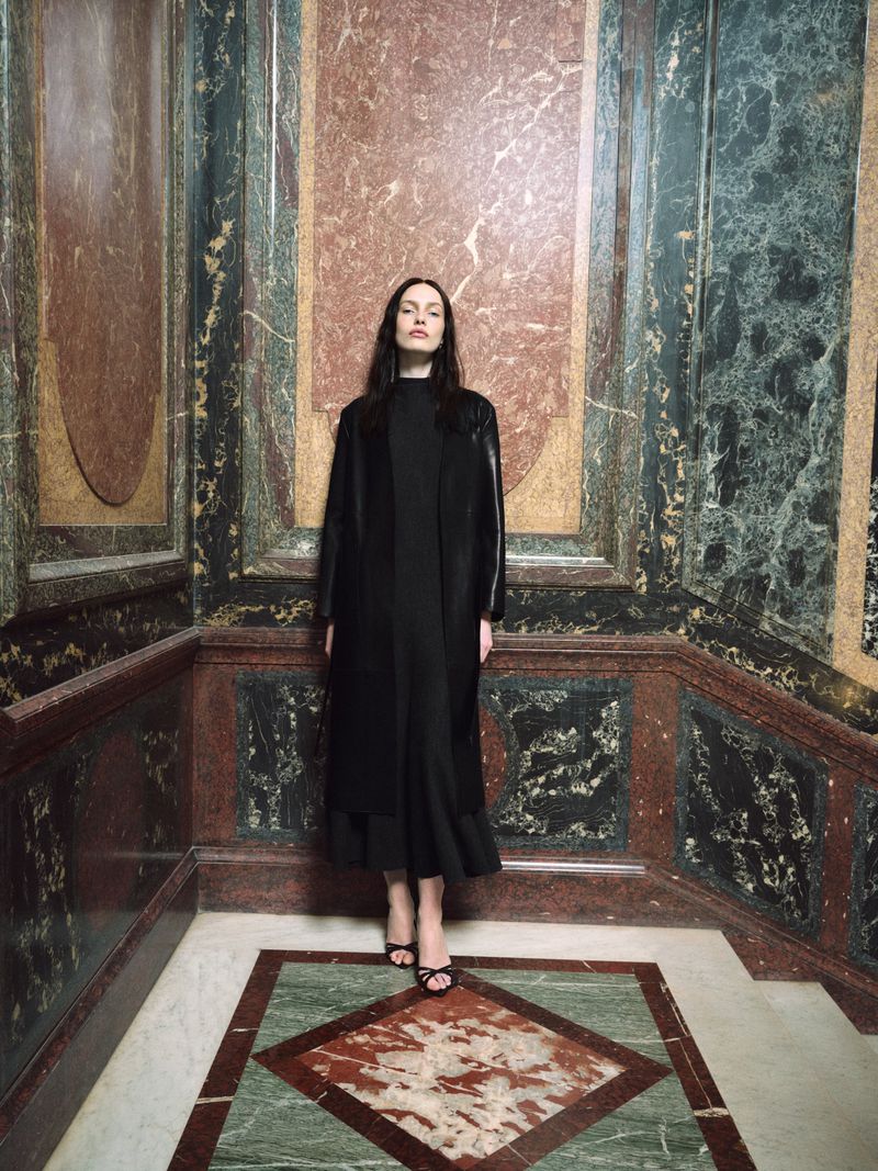 Model in a black coat standing against ornate marble and stone walls for Nour Hammour Summer 26 campaign.