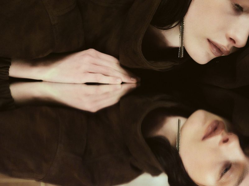 Close-up portrait of a woman reflected in a mirror wearing a brown jacket with crystal earrings.