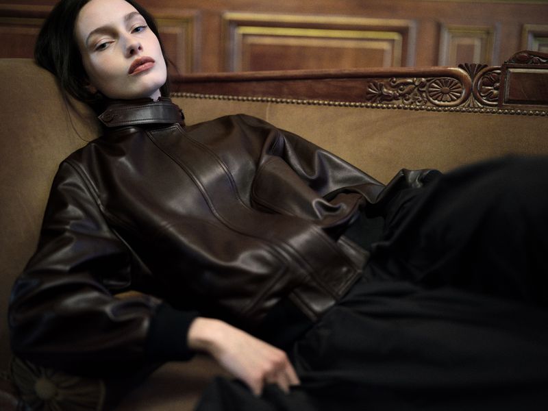 Model reclining on an antique couch wearing a dark brown leather jacket against a wood panelled background.