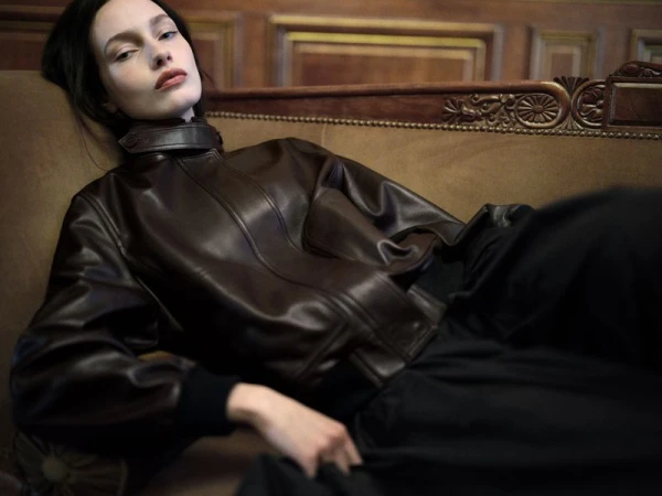 Model reclining on an antique couch wearing a dark brown leather jacket against a wood panelled background.