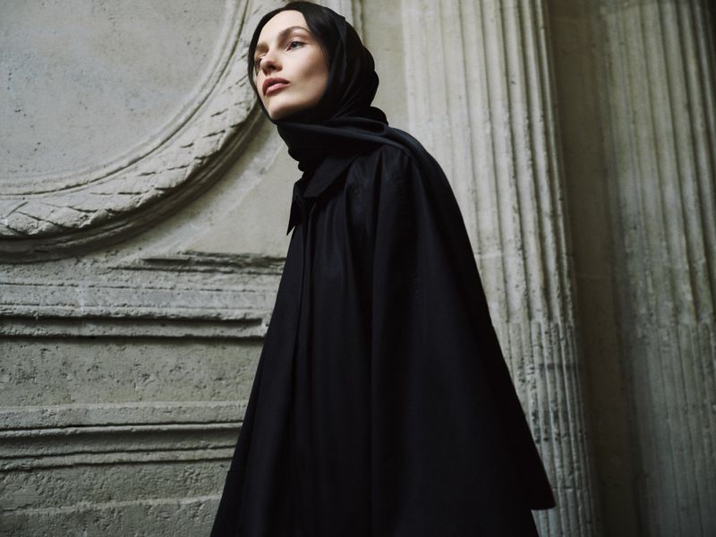 Model wearing a dark headscarf and black cape posing against classical stone architectural pillars.