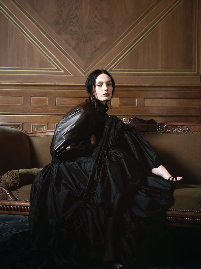 Model wearing a dark leather jacket and voluminous black gown seated on a brown sofa against wooden paneling.
