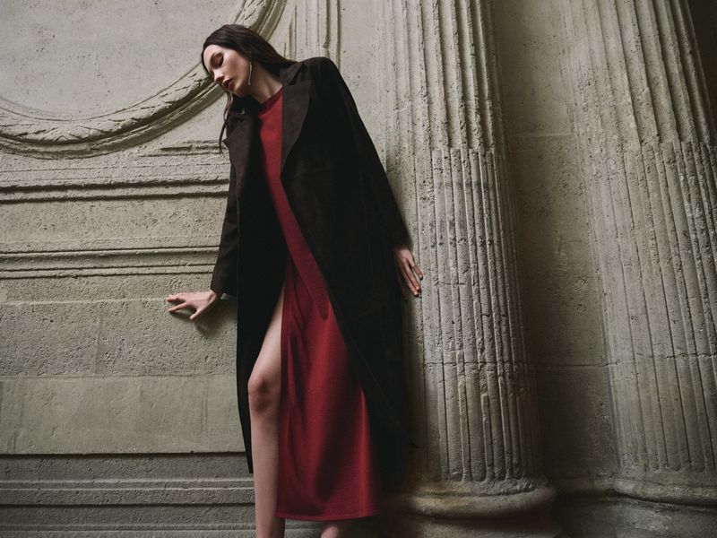 Model in long dark coat wearing a red dress leaning against neoclassical stone pillars for Nour Hammour Summer 26