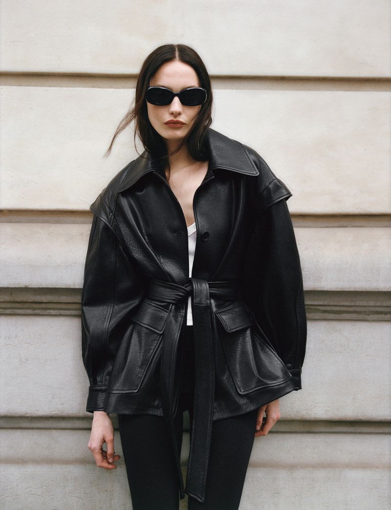 Model in dark sunglasses and a black leather belted jacket posing against an architectural stone wall exterior.