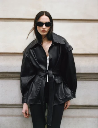 Model in dark sunglasses and a black leather belted jacket posing against an architectural stone wall exterior.