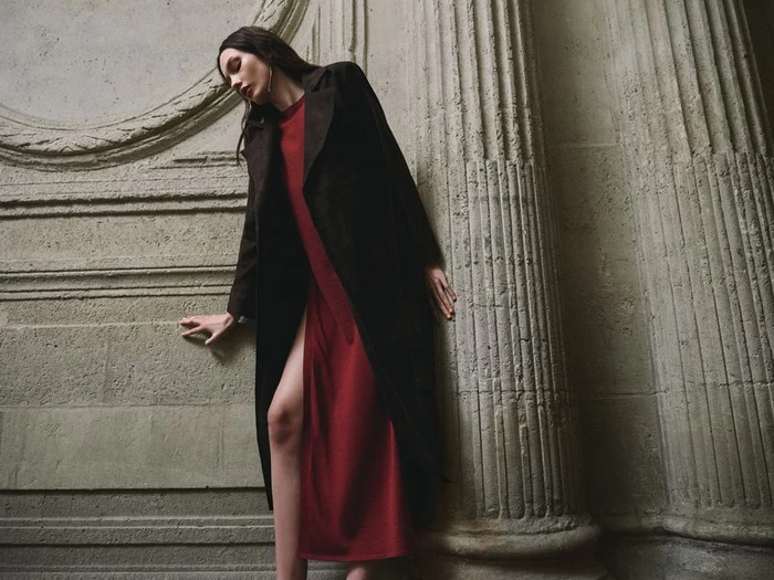 Model in long dark coat wearing a red dress leaning against neoclassical stone pillars for Nour Hammour Summer 26