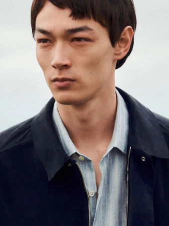 Close-up portrait of a man wearing a dark jacket over a light striped collared shirt outdoors against an overcast sky.