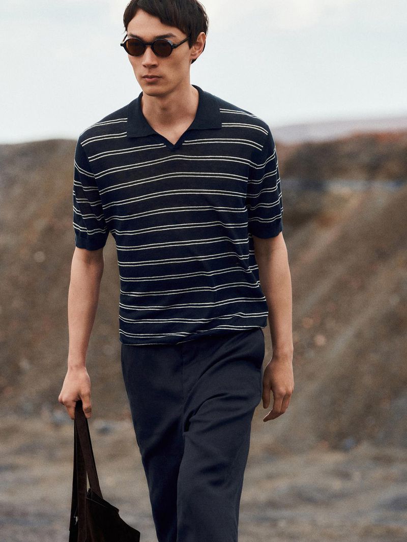 Model in dark sunglasses and striped polo shirt walking outdoors against a terrain background for Massimo Dutti