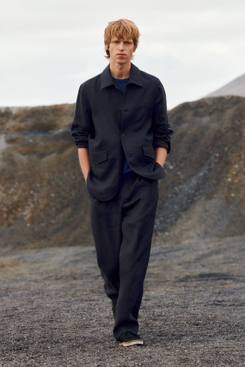 A male model wearing a dark button-down shirt and matching trousers walks along a dark rocky path under an overcast sky.
