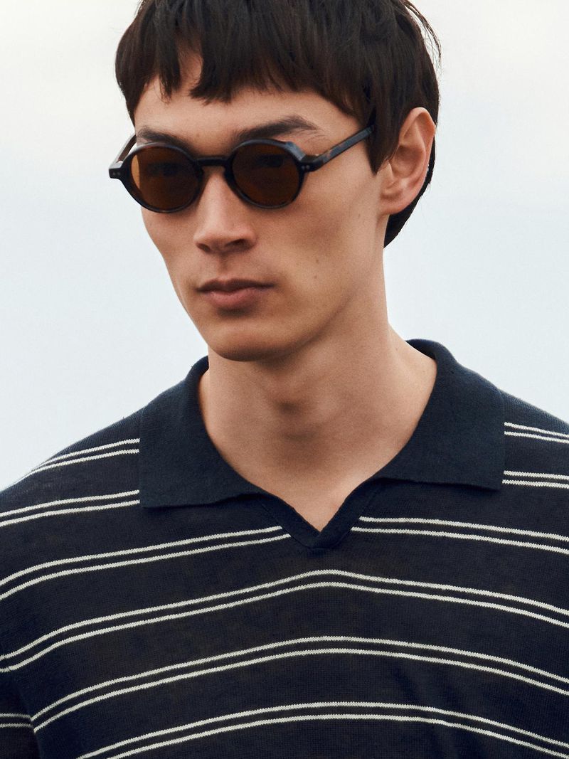 Man wearing round dark sunglasses and a black-and-white striped polo shirt against an overcast sky.