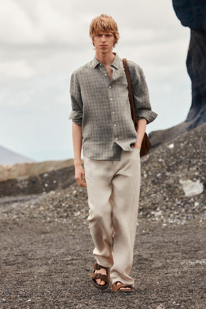 Model walking outdoors on dark volcanic sand wearing a grey patterned linen shirt and light beige trousers