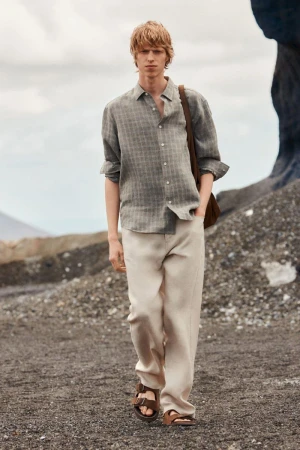 Model walking outdoors on dark volcanic sand wearing a grey patterned linen shirt and light beige trousers