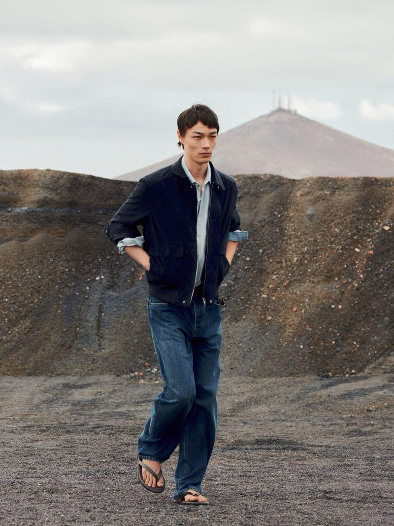 Model walking across a rugged terrain wearing a dark jacket, blue shirt, and jeans under an overcast sky.