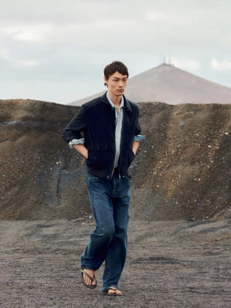 Model walking across a rugged terrain wearing a dark jacket, blue shirt, and jeans under an overcast sky.