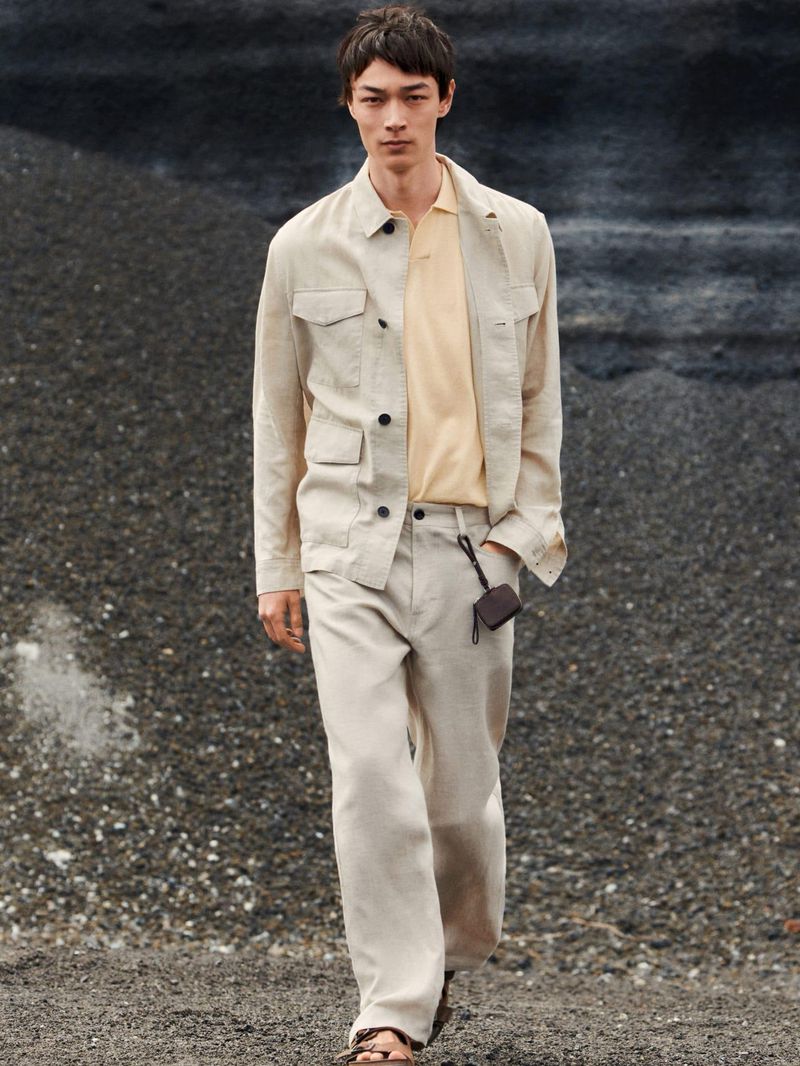 Male model wearing a light beige linen utility jacket and trousers over a pale yellow polo shirt against a rocky terrain background.