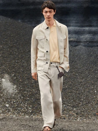 Male model wearing a light beige linen utility jacket and trousers over a pale yellow polo shirt against a rocky terrain background.