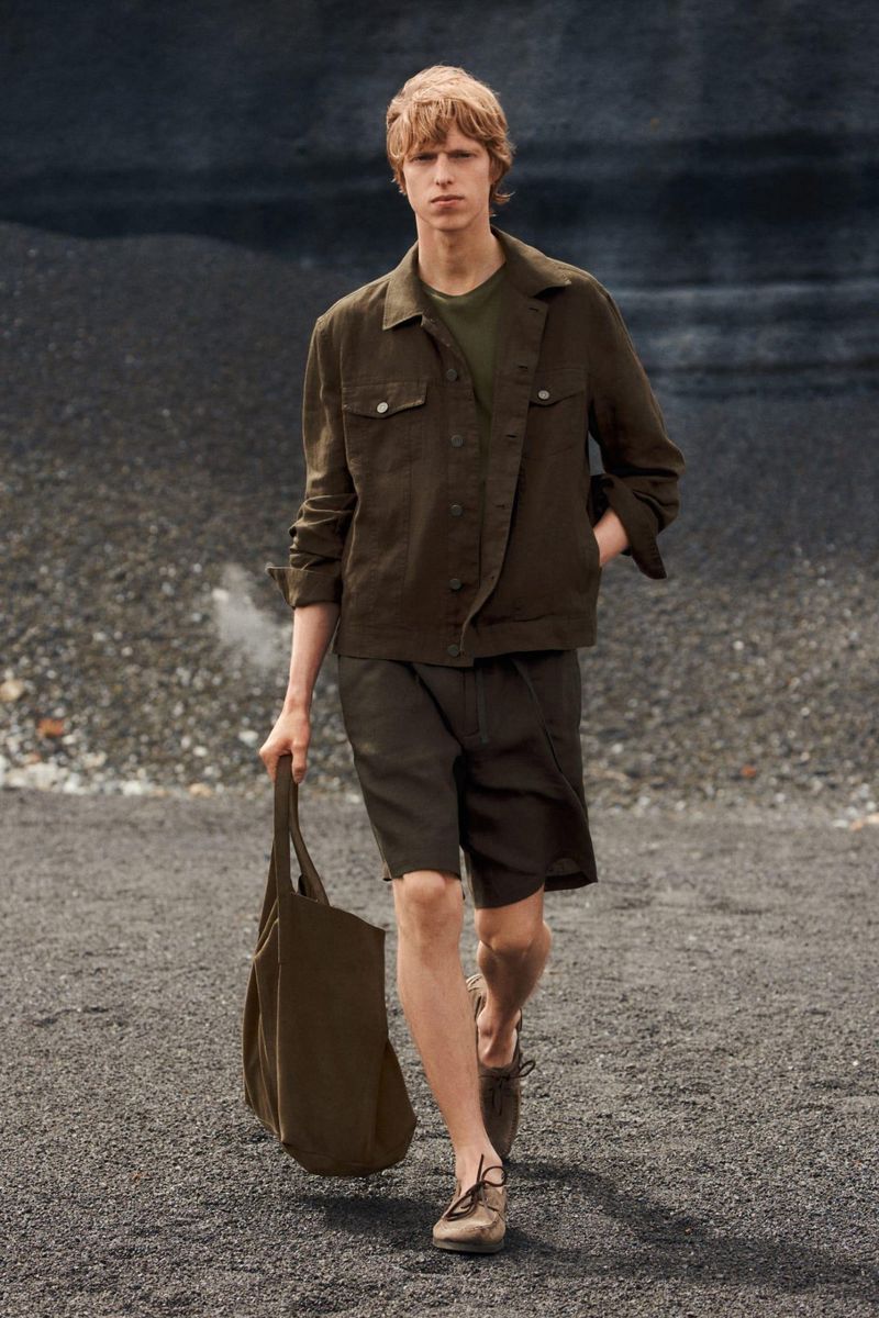 Male model in an olive green jacket and shorts walking on dark sand holding a matching tote bag