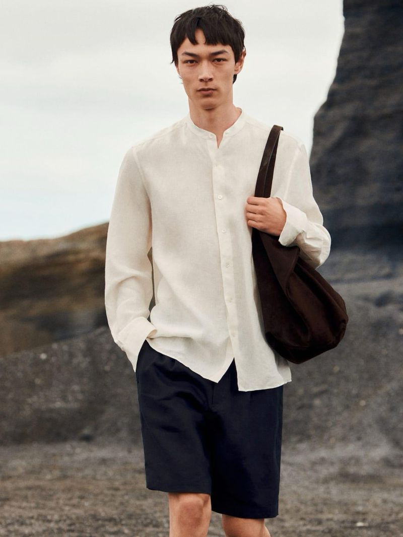 Model in white long-sleeve shirt and dark shorts carrying a brown shoulder bag on rocky terrain