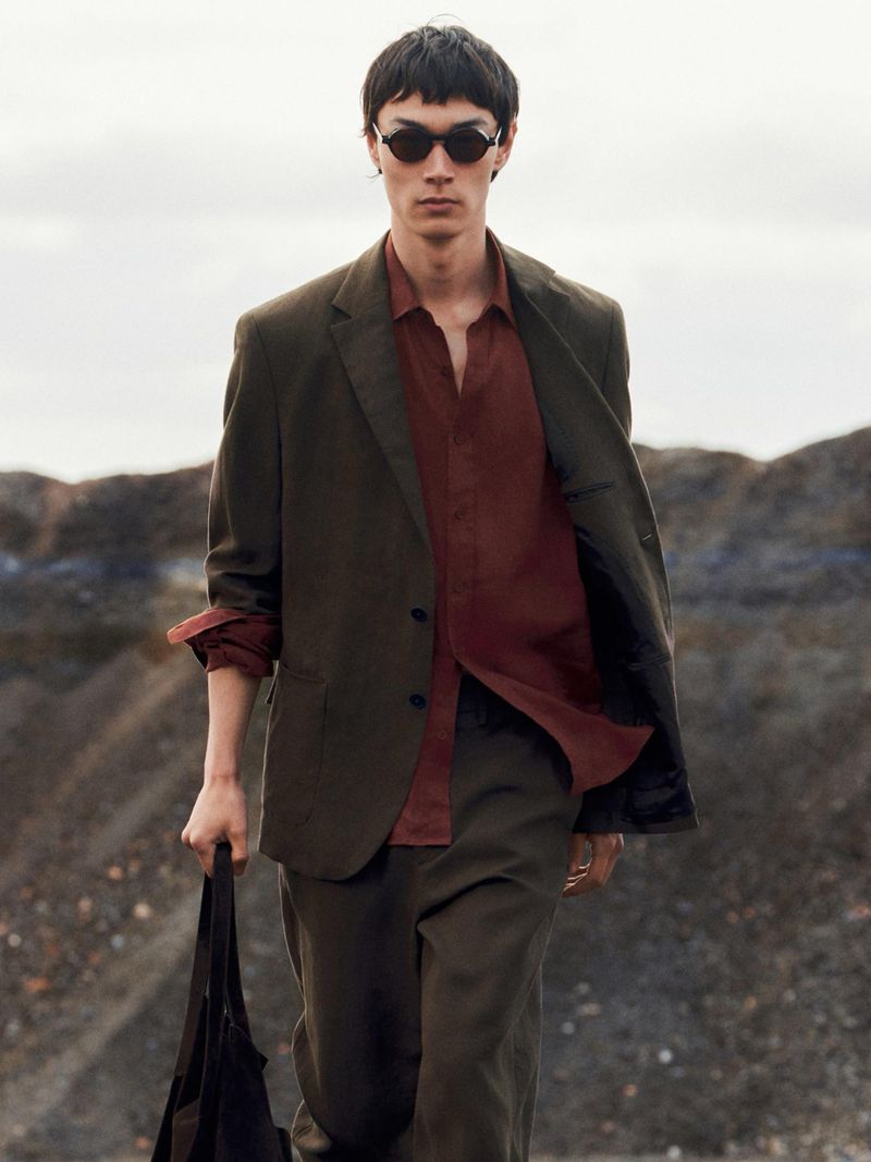 Model in dark sunglasses wearing an olive suit jacket and rusty red shirt holding a black bag outdoors against a terrain background