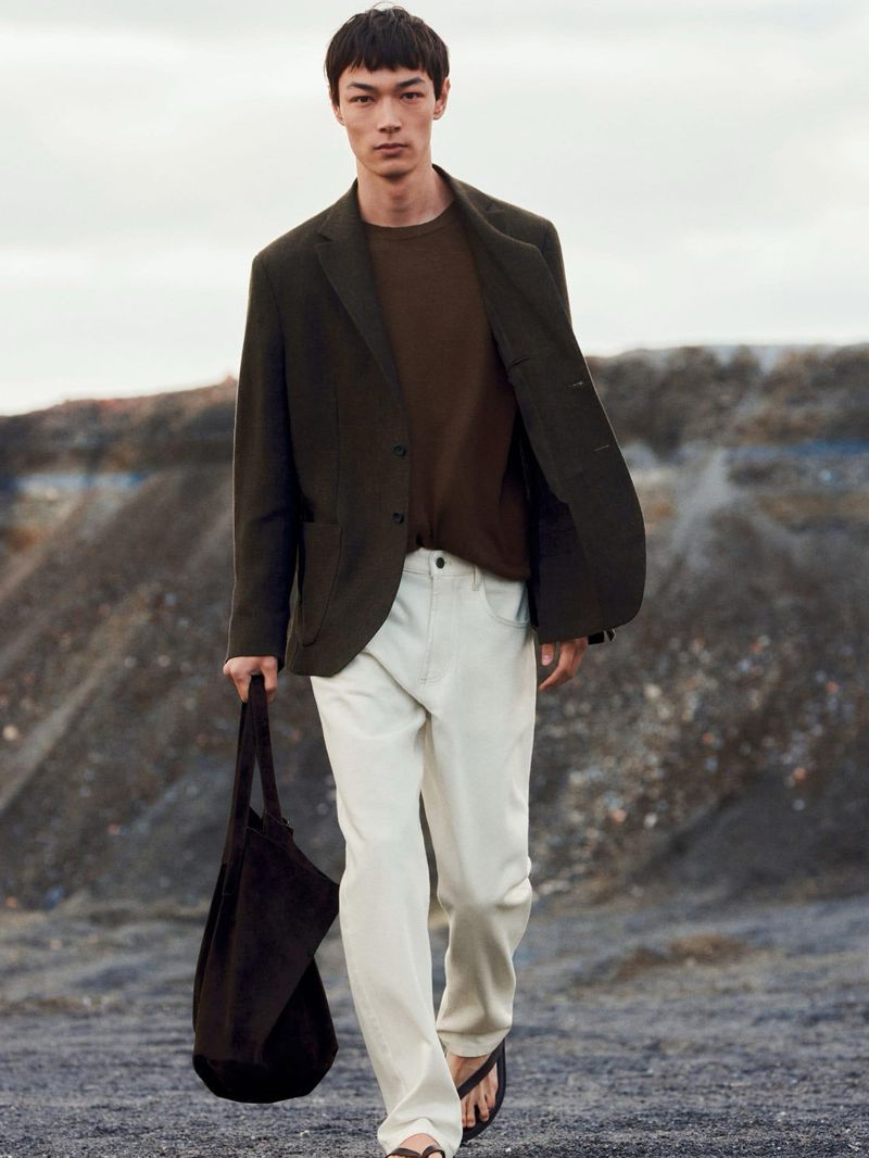 Male model in an olive blazer and white trousers walking outdoors carrying a dark tote bag against a rugged terrain background