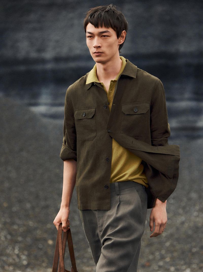 Model wearing an olive green overshirt over a yellow polo shirt holding a bag outdoors