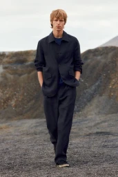 A male model wearing a dark button-down shirt and matching trousers walks along a dark rocky path under an overcast sky.