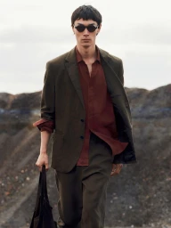 Model in dark sunglasses wearing an olive suit jacket and rusty red shirt holding a black bag outdoors against a terrain background
