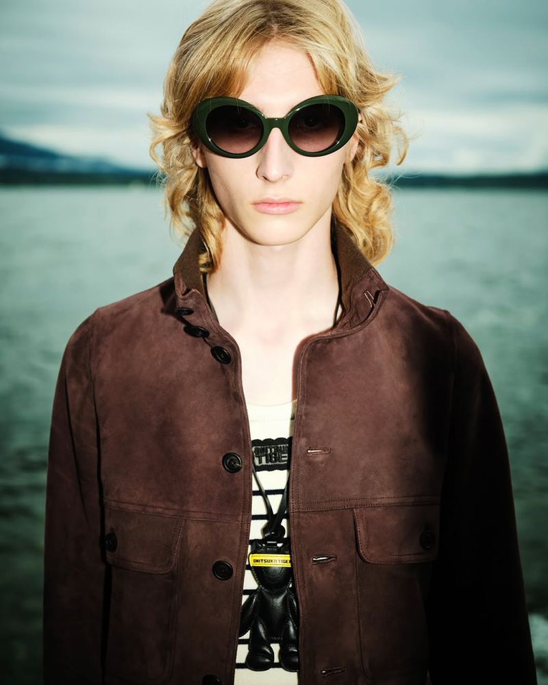 Close-up portrait of a blonde model wearing dark oversized sunglasses and a brown jacket with a lake background.