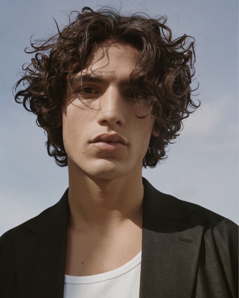 Portrait of a young male model with dark curly hair wearing a black jacket and white tank top against a clear sky background
