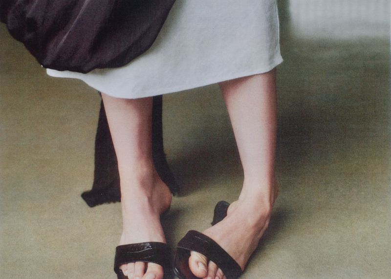 Close-up of a model wearing black sandals, a white skirt, and a dark garment against a neutral floor background.