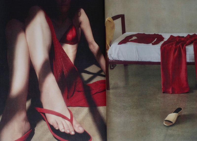 Model seated wearing red top and sandals next to a bed with red clothing in an editorial interior setting