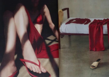 Model seated wearing red top and sandals next to a bed with red clothing in an editorial interior setting