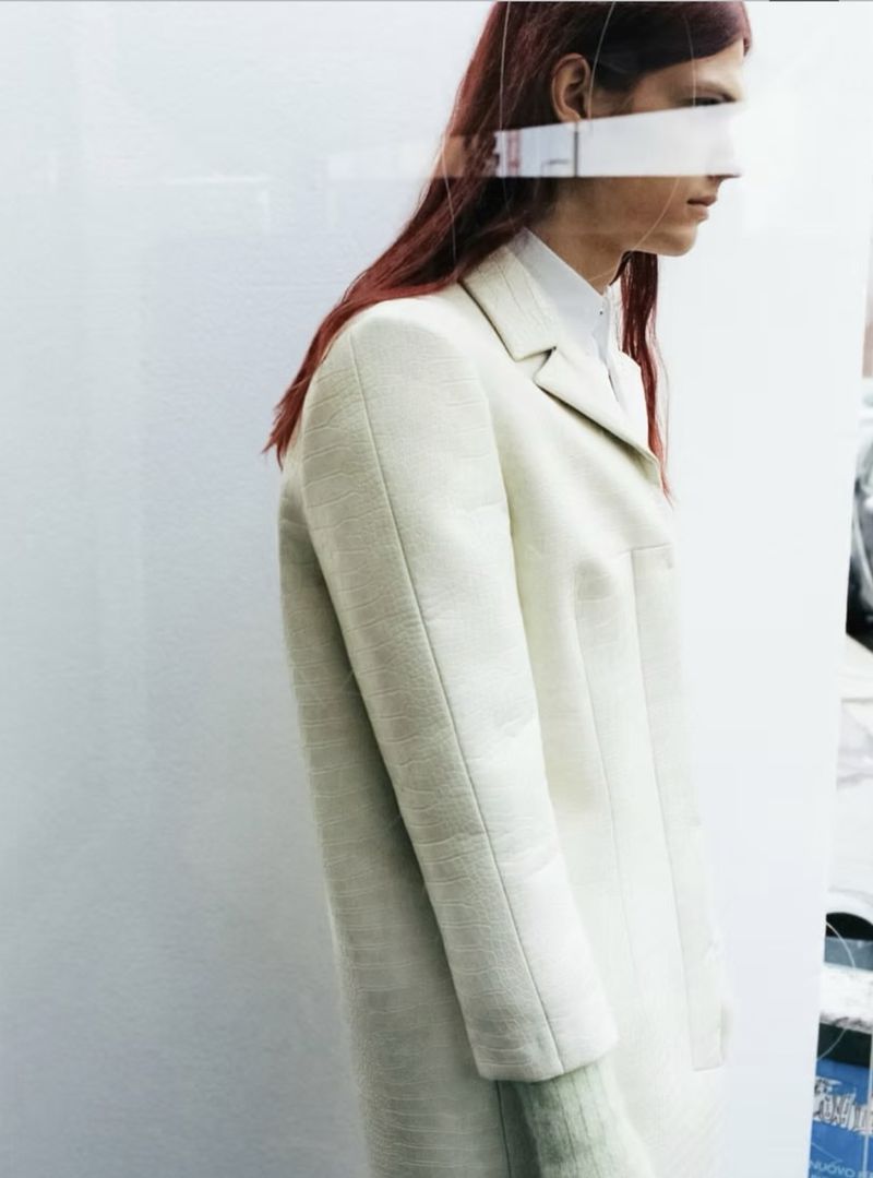 Model with long reddish hair wearing an off-white coat against a plain background