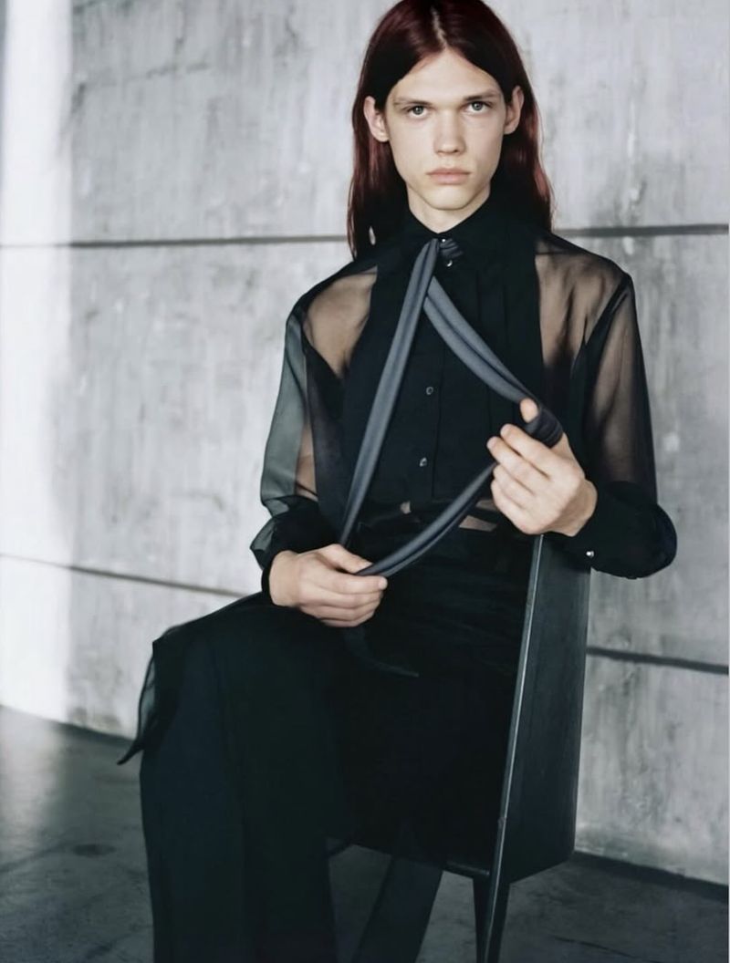 Model with long reddish-brown hair seated in a dark chair wearing a sheer black shirt holding grey straps against a concrete wall