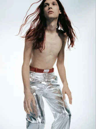 Shirtless male model with long dark hair wearing metallic silver trousers against a white background