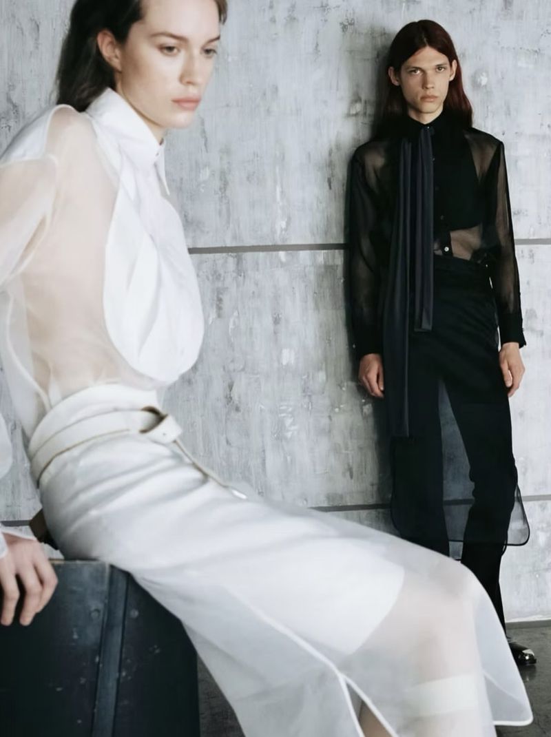 Woman in white sheer blouse and skirt seated in foreground with man in black sheer shirt standing behind against a textured concrete wall