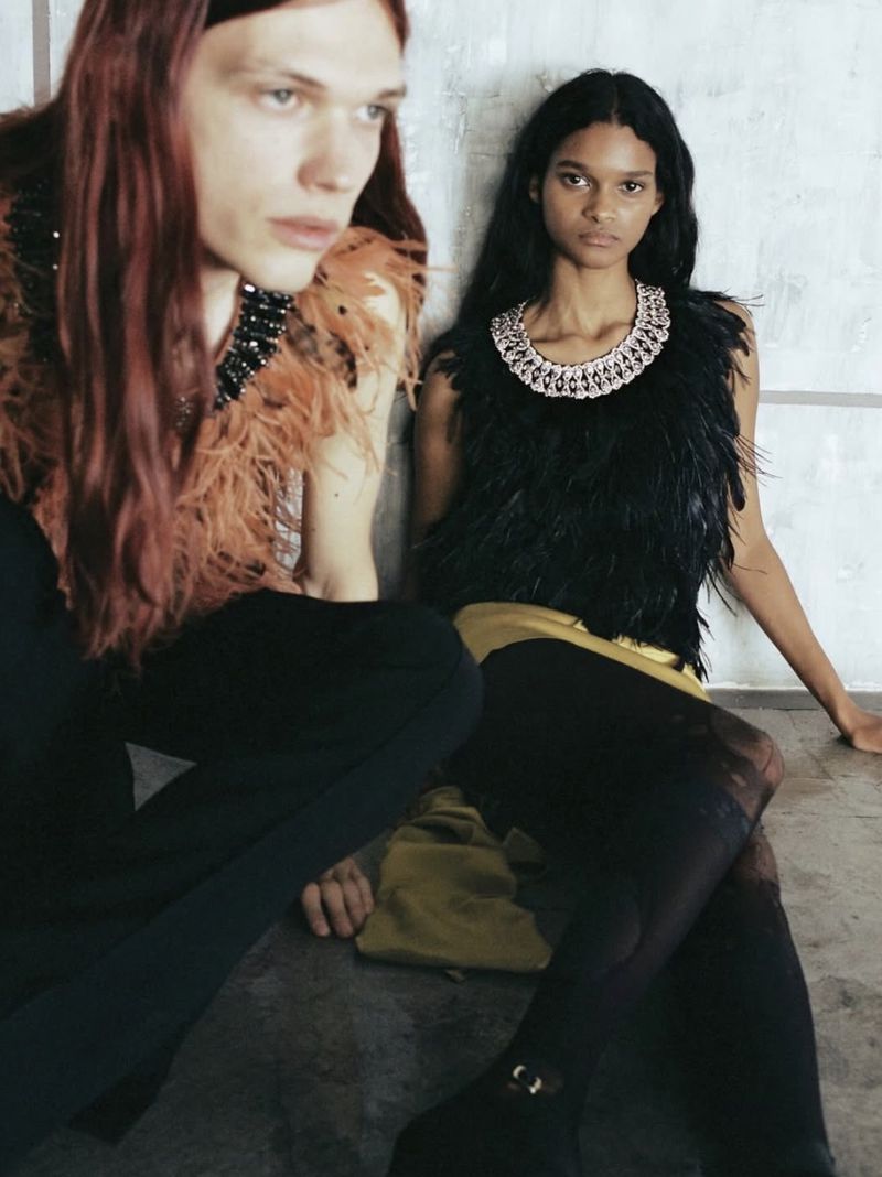 Two models posing indoors against a textured wall, wearing feathered sleeveless tops and ornate jeweled collars.