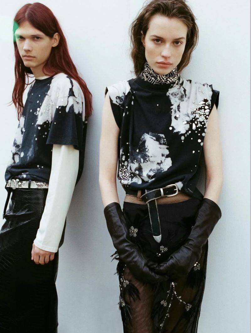 Two models wearing patterned black and white tops and dark skirts pose against a plain light wall in a fashion editorial.