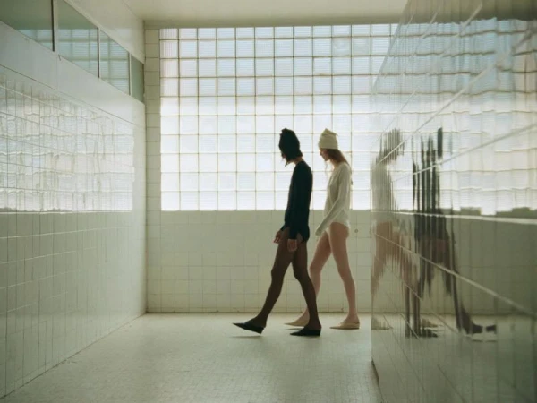 Two slim adults walking in a minimalist, tiled interior wearing simple leotards and beanies, illuminated by bright window light.