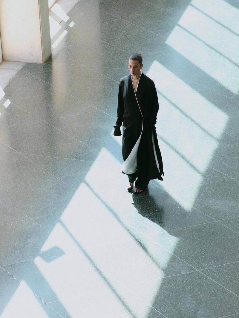 Model in long black coat walking across a large grey stone hall with strong diagonal light bars