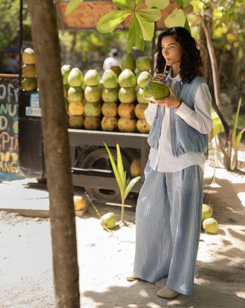Model drinking from a fresh coconut in a blue striped vest and matching pants outdoors near a coconut cart