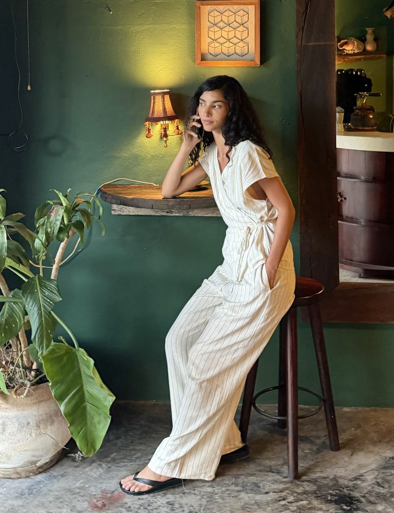Model seated on a wooden stool against a dark green wall, wearing a striped white jumpsuit and making a phone call.