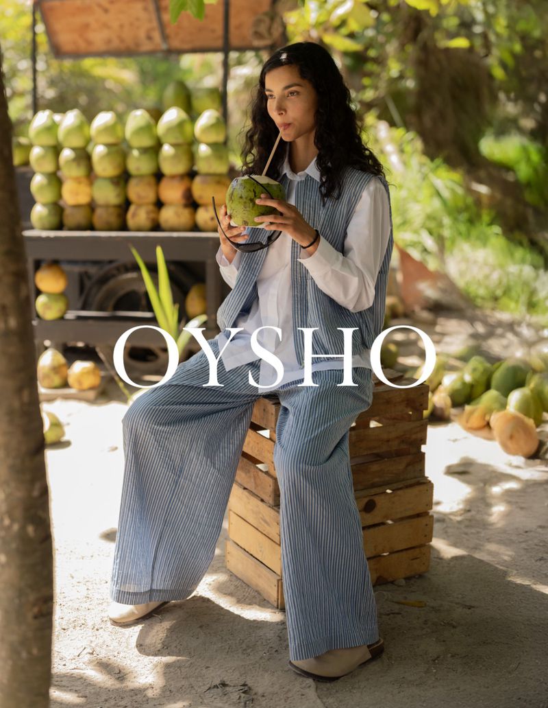 Model drinking from a fresh coconut while wearing a blue striped knit vest and matching wide-leg trousers outdoors near a fruit cart.