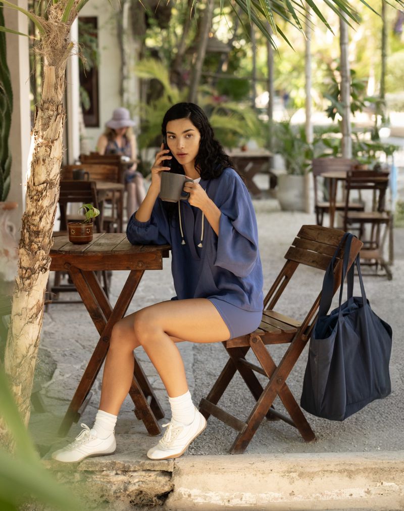 Woman in blue hoodie and shorts seated at a wooden cafe table holding a mug and phone, with a dark tote bag on a chair.