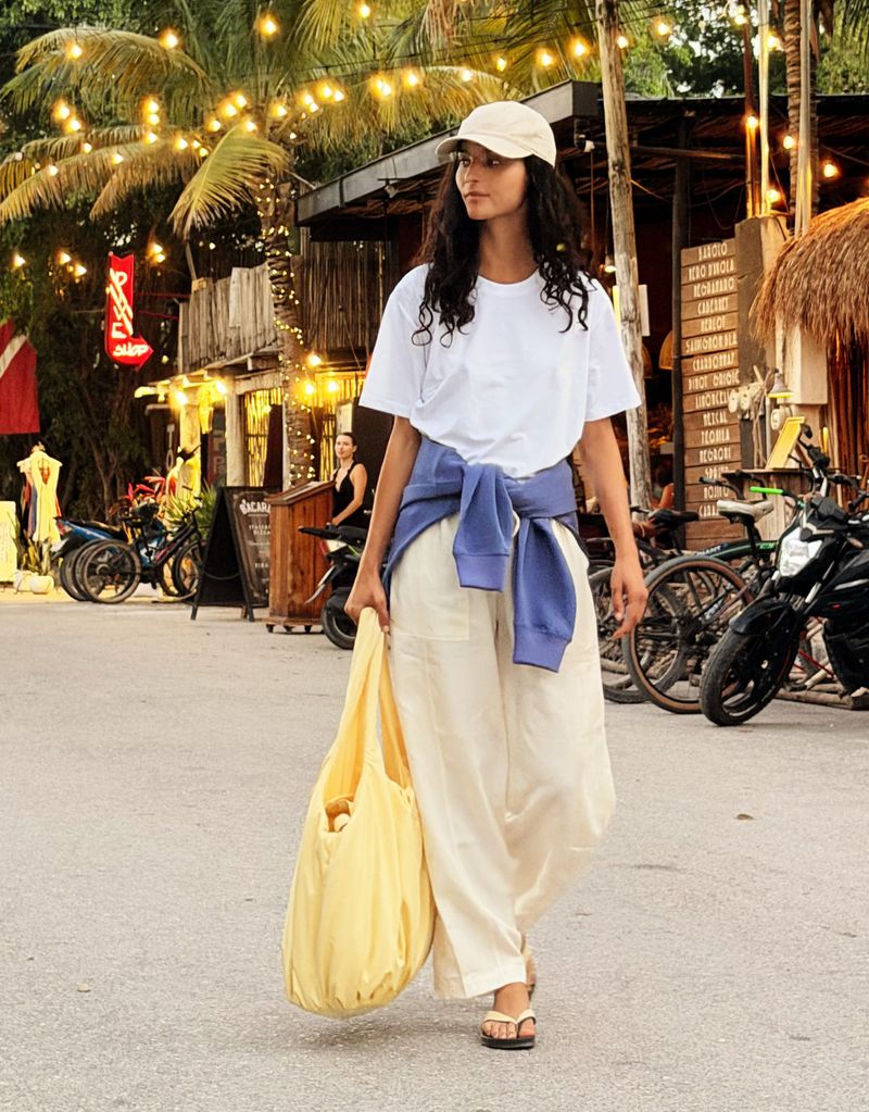 Woman in casual clothes walking on a paved road with palm trees and buildings in the background, carrying a yellow tote bag.