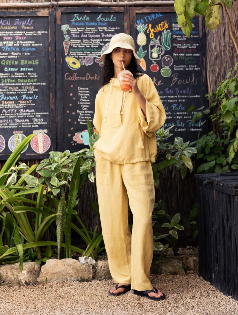 Model Tindi Mar wearing a yellow linen hoodie and pants holding a smoothie in front of a cafe menu board for Oysho