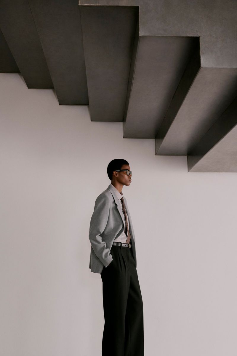 Model wearing a grey blazer and black trousers standing below sculptural concrete stairs against a plain wall